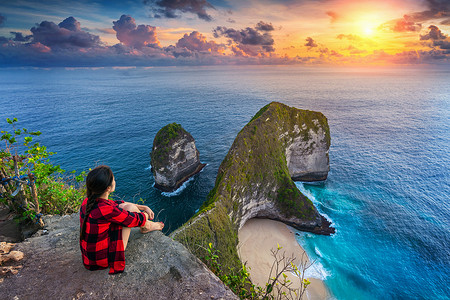 努沙摄影照片_印度尼西亚巴厘岛努沙佩尼达岛的 Kelingking 海滩上，一名妇女坐在悬崖上欣赏日落。
