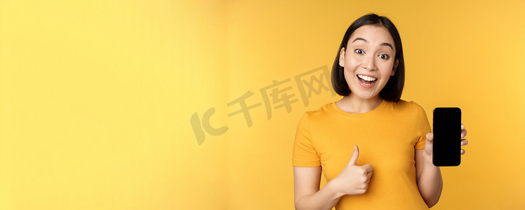 兴奋的亚洲女性展示手机屏幕，竖起大拇指，喜欢某事，推荐智能手机应用程序，站在黄色背景上