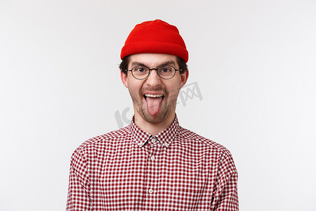 特写肖像滑稽可爱的留着胡子的嬉皮士，戴着红色的帽子和眼镜，笑着开玩笑地吐舌头，玩得开心，表现得像个孩子气的站在白色背景上的快乐