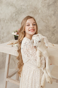女孩抱着花摄影照片_女孩坐在复活节餐桌旁，手里拿着蛋糕、春花和鹌鹑蛋，怀里抱着一个白人山羊。