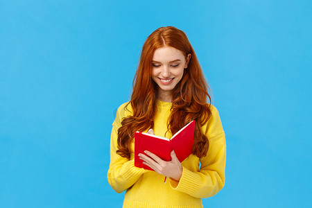 腰上肖像红发女学生在红色可爱的笔记本上写下想法，准备购物清单，为大学讲座制定时间表，微笑着在日记中分享想法，蓝色背景