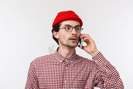 特写：戴眼镜、头戴红帽的年轻白人男子交谈、打电话给朋友四处寻找人群中的人、将智能手机放在耳朵附近、白色背景