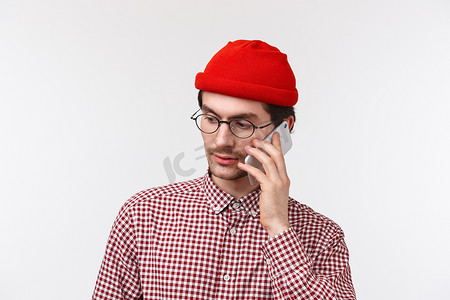 确认订单摄影照片_特写：穿着红色帽子和眼镜的严肃年轻人用手机打电话给某人，交谈，在下订单或确认预订时侧视，白色背景