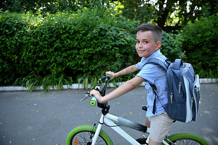 可爱的小男孩背着书包，骑着自行车可爱地笑着看着镜头。