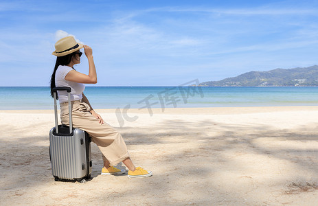 夏天，年轻快乐的女人坐在沙滩上的旅行箱上。
