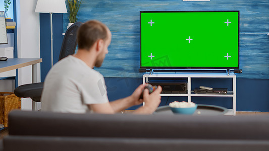 男子坐在沙发上拿着无线控制器在绿屏电视上玩控制台视频游戏