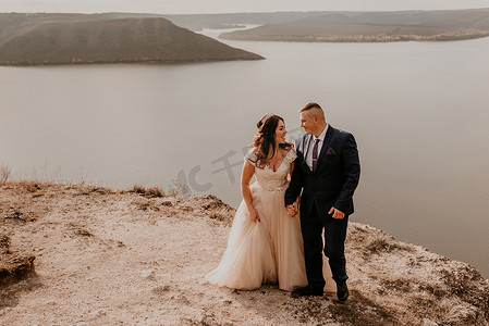 礼服首饰摄影照片_夏天，一对穿着白色礼服和西装的情侣在河上的山上举行婚礼