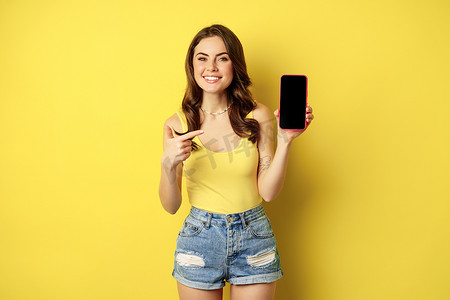 年轻时尚女性展示手机屏幕，指着智能手机显示屏、应用程序界面或在线商店，微笑着高兴，站在黄色背景上