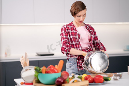 蔬菜清洗摄影照片_年轻女子清洗或混合金属锅中的配料和柜台上的蔬菜。