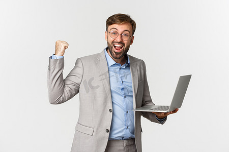 穿着西装和眼镜的快乐商人的肖像，拿着笔记本电脑，满意地微笑着，说是，并做出拳头泵的手势，在工作中实现目标，站在白色背景上