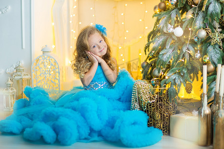 除夕夜背景摄影照片_年轻漂亮的女孩穿着蓝色白色优雅的晚礼服，坐在圣诞树附近的地板上，在除夕夜送礼物。