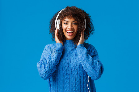 快乐迷人的非洲裔美国微笑女孩得到圣诞礼物新耳机，听音乐，享受美妙的节拍，触摸耳机，看着相机，蓝色背景