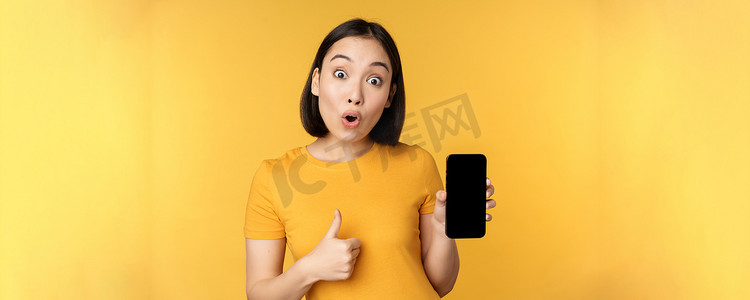 兴奋的亚洲女性展示手机屏幕，竖起大拇指，喜欢 smth 好，推荐智能手机应用程序，站在黄色背景上