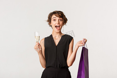 很奇怪摄影照片_兴奋的时尚女性看起来很奇怪，拿着购物袋和一杯香槟，站在白色背景上庆祝派对