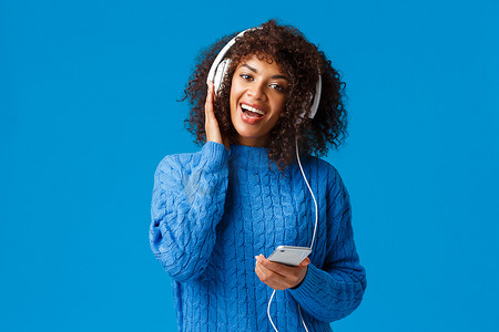 快乐开朗的非洲裔美国女性在智能手机上玩卡拉 OK 游戏应用程序，戴着耳机唱歌，享受美妙的音乐、音质、站立的蓝色背景
