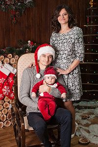 一个家庭带着一个穿着圣诞老人服装的小孩在家里坐在摇椅上的壁炉前。