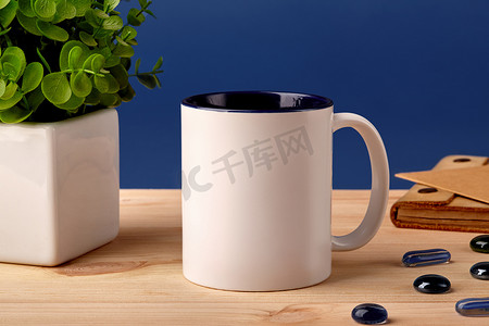 石子摄影照片_木制桌面上的白色陶瓷杯，旁边是散落的玻璃石、盆中的绿色植物和棕色记事本，蓝色背景。