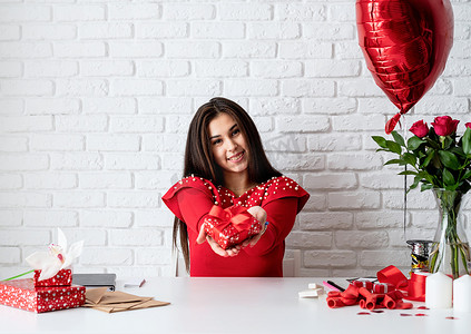 白红背景摄影照片_白砖墙背景下，身穿红裙的年轻浪漫女性拿着情人节礼物