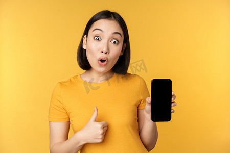 兴奋的亚洲女性展示手机屏幕，竖起大拇指，喜欢 smth 好，推荐智能手机应用程序，站在黄色背景上