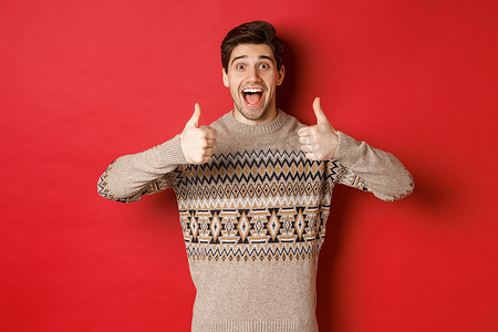 穿着圣诞毛衣的快乐男人的画像，就像一件好事，竖起大拇指，微笑着惊讶，赞美新年派对，站在红色背景上