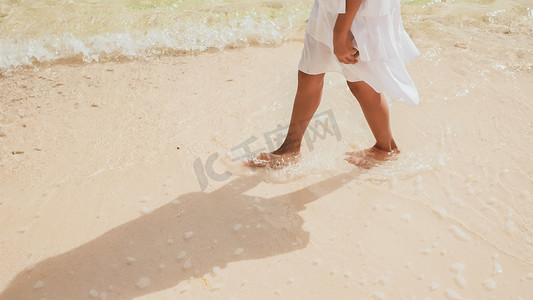 晒水摄影照片_身穿白色连衣裙的菲律宾女学生晒黑的双腿沿着白色的沙滩奔跑，触碰着沙滩上的浪花。