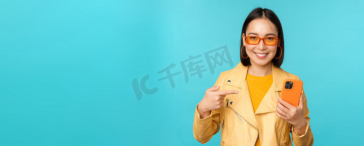 年轻时尚的亚洲女模特戴着时髦的太阳镜、春装、展示手机、指着智能手机微笑，站在蓝色背景上