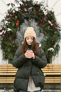 女人在城市街道上喝热饮，背景是咖啡馆的窗户，上面装饰着圣诞花环