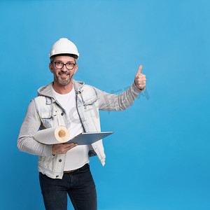 中年英俊的建筑男子拿着一卷壁纸和数字平板电脑，穿着时尚的休闲牛仔服，戴着安全帽或头盔，竖起大拇指或在蓝色背景上完美隔离