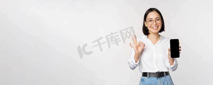 亚洲女商人的形象，显示智能手机屏幕、应用程序界面和 ok 标志，推荐手机上的应用程序，站在白色背景上