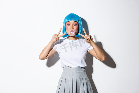 戴着蓝色假发的愚蠢的亚洲女孩，打扮成动漫人物，化着五颜六色的妆，表现出和平的姿态，吹着泡泡糖，站在白色背景上