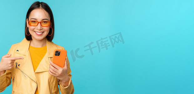 春装尚新摄影照片_年轻时尚的亚洲女模特戴着时髦的太阳镜、春装、展示手机、指着智能手机微笑，站在蓝色背景上