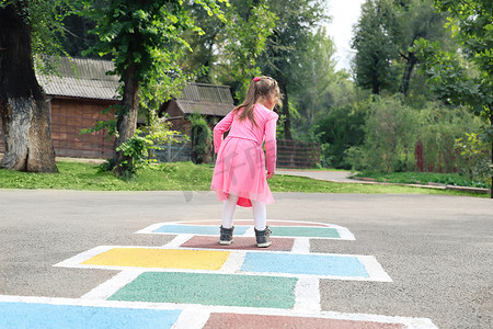 跳房子摄影照片_穿着粉红色连衣裙的小女孩在街上玩跳房子。