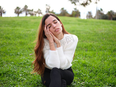 公园里一位年轻女子坐在草地上，双手捂着脸，闭着眼睛做白日梦。
