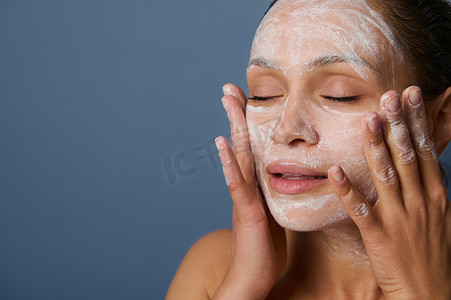 美丽的女人在用泡沫清洁化妆品卸妆的同时按摩她的脸，并用一种​​在灰色背景下隔离的去角质美容产品使她的皮肤焕然一新。