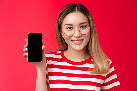 特写可爱的亚洲金发女孩推广智能手机应用程序，显示个人社交媒体页面的女博主拿着手机看着相机开心地微笑，推荐设备吹嘘自己的分数，红色背景