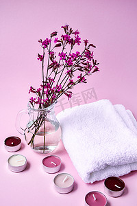 粉色花朵小花朵摄影照片_白色棉毛巾，粉色背景上有小粉色花朵和芳香蜡烛，水疗概念，复制空间