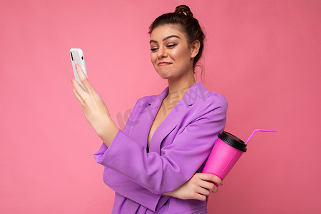 饮料照片摄影照片_一张快乐大笑的白人黑发女人拿着外带咖啡的照片，穿着休闲服，隔离在粉色墙壁上，使用智能手机