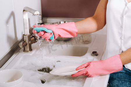 戴粉红色手套的家庭主妇女孩用洗涤剂在水槽里用手洗碗。