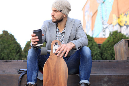 英俊的年轻人穿着灰色外套和帽子，休息，坐在长板上喝咖啡。