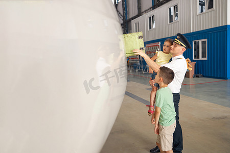 身着制服的专业飞行员向他的两个小孩展示飞机的部件，他们来飞机库看望他们的父亲