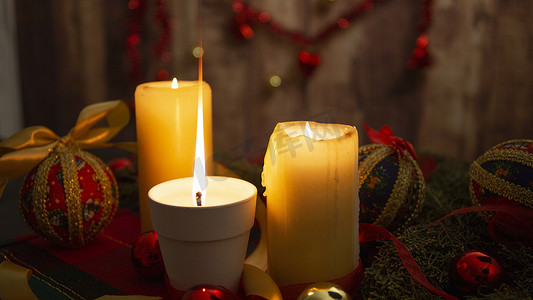 圣诞桌布上点燃的大火焰蜡烛的特写，周围有松树枝、剪纸小饰品、点燃的蜡烛和悬挂在木制背景上的圣诞装饰，具有散景效果