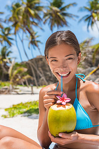 美丽的亚洲比基尼女人直接从海滩上的稻草水果中喝新鲜椰子水。