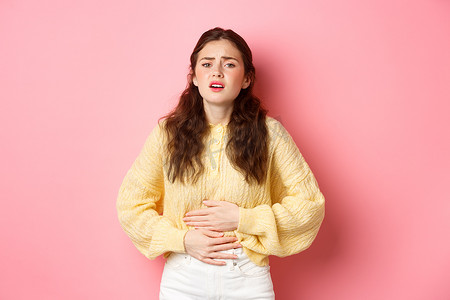 胃痛的年轻女性，双手放在肚子上，感觉非常疼痛，月经来潮，站在粉红色的背景下
