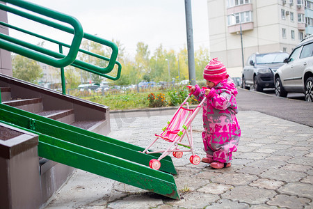 带着玩具婴儿车的可爱蹒跚学步的孩子沿着轮椅、手推车和婴儿车的钢栏杆坡道行走。