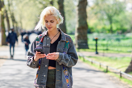 现代发型的年轻都市女性使用智能手机走在城市公园的街道上。