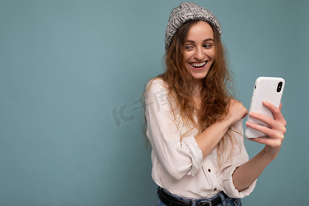 蓝色背景墙上隔着蓝色背景墙，身穿休闲白衬衫和灰色帽子、手持电话、使用在线交流的漂亮年轻卷发女性的照片
