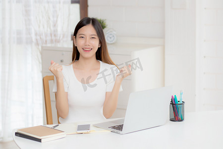 年轻的亚洲女性自信地在家里用笔记本电脑工作，思考规划的想法，自由职业的快乐女孩兴奋并成功地使用笔记本深思熟虑和写作，商业理念。