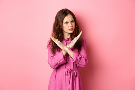 愤怒的女人阻止提议，表现出交叉停止手势，说不，摇头否定回答，站在粉红色背景上