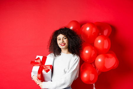 美丽的白人女性，深色卷发，红唇，拿着浪漫的心形气球站着，放在盒子里，收到情人的礼物，站在工作室的背景上