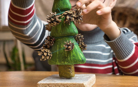 创意圣诞节摄影照片_一个小男孩用松果装饰一棵手工制作的圣诞树，同时玩耍。